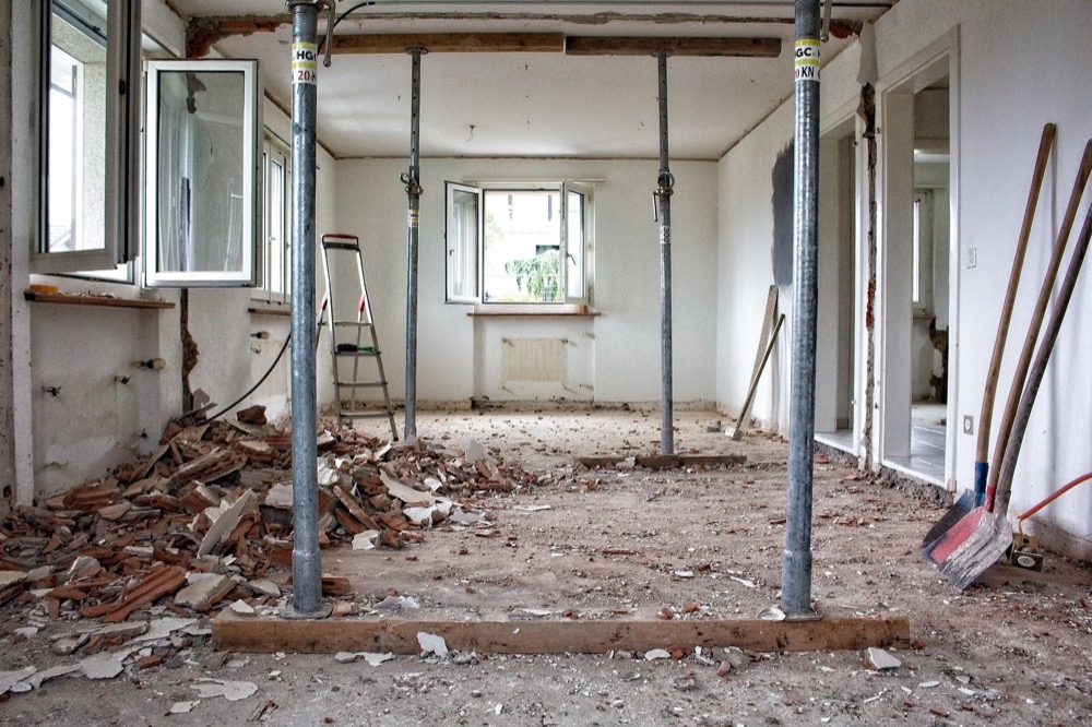 Baustelle: Raum mit Schutt, vier Stützpfosten, zwei offene Fenster, zwei Schaufeln und Besen an der Wand.