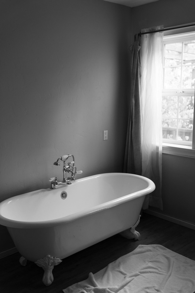 Schwarz-weiße Ansicht einer freistehenden Badewanne mit Klauenfüßen, Holzboden und Fenster mit Vorhängen.