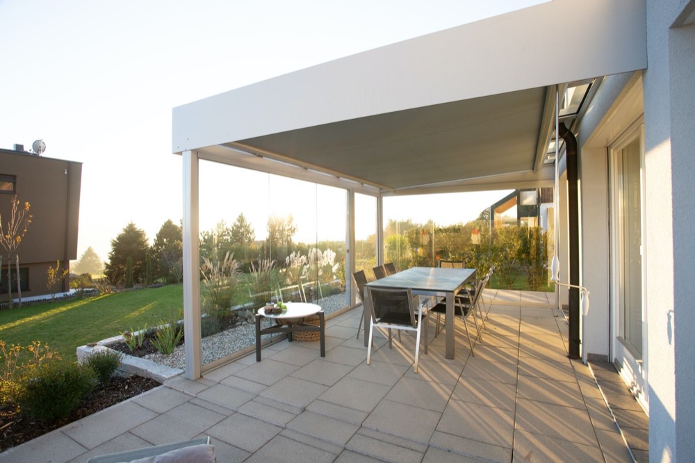Moderne Terrasse mit grauer Pergola, Glastisch und Stühlen vor einem Haus mit Garten bei Sonnenuntergang.