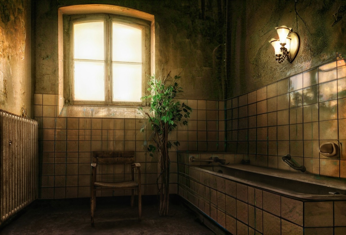 Dunkles, altes Badezimmer mit Fenster, Holzstuhl, Pflanze und Badewanne mit Fliesen.